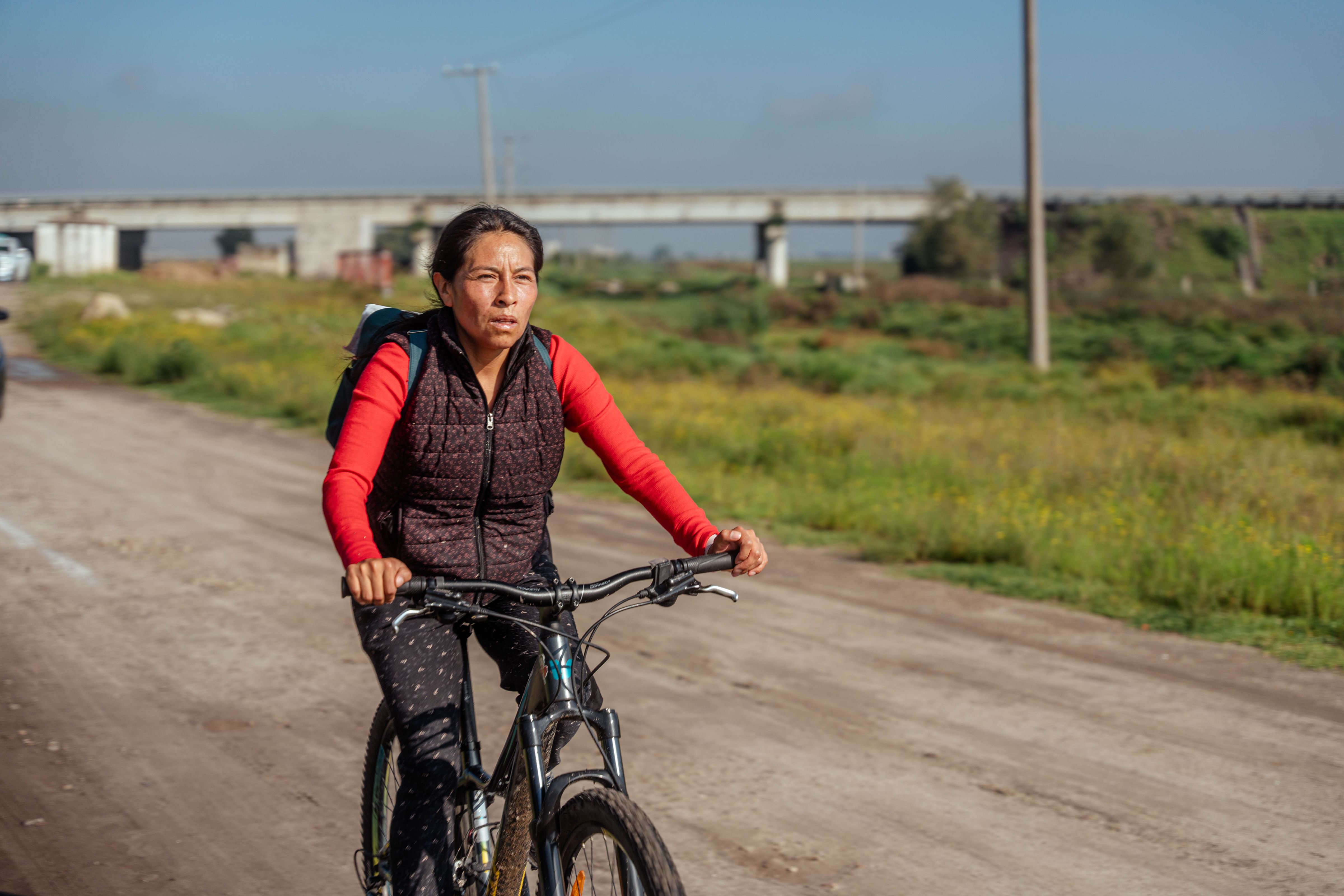 A woman rides a mountain bike toward the camera on a dusty dirt road, fields open behind her.